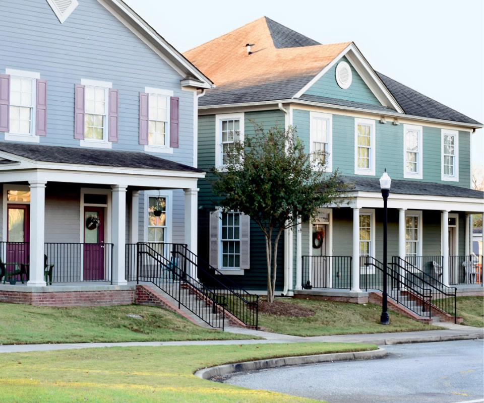 A suburban street with two adjacent, two-story houses featuring front porches and well-manicured lawns. One house is painted blue with burgundy shutters, and the other is green with white trim. A tree and streetlamp are visible in the foreground.