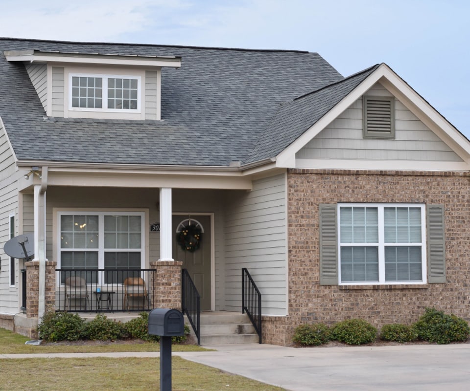 A two-story house with a sloped gray roof and a mix of brick and light-colored siding. The front porch has a small seating area with two chairs and a table. There's a wreath on the front door, and a mailbox is in the foreground.