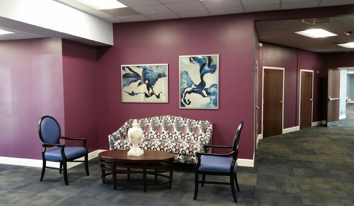 A waiting area with purple walls features a patterned sofa, two blue chairs, and a wooden coffee table. Two abstract paintings with blue and white swirls hang above the sofa. A white decorative bust is on the table. Doors are visible in the background.