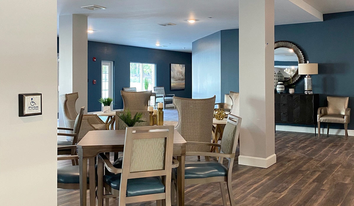 A modern, spacious room with a dining area featuring a wooden table and chairs. In the background, there are armchairs near large windows. The walls are painted dark blue, and a circular mirror decorates the far wall. Lighting is soft and inviting.