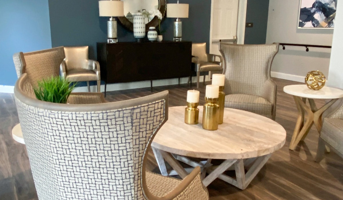 Modern lounge area with woven chairs, a round wooden table, and decorative candles. A console table with lamps and vases is against a teal wall. Two metallic chairs and wall art complete the sophisticated decor.