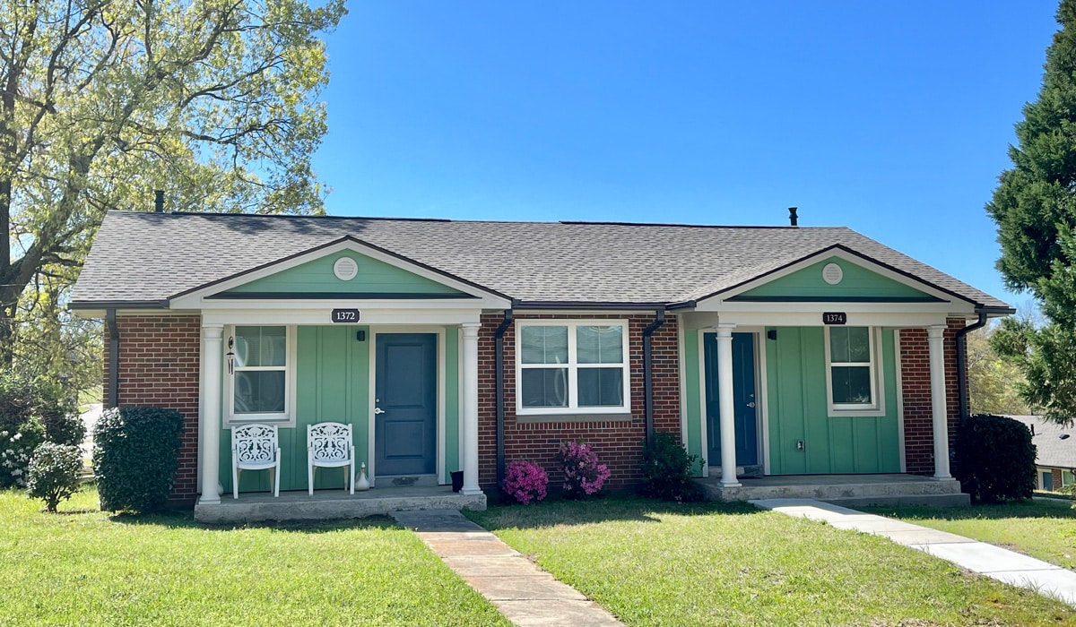 A small brick duplex with a gable roof, featuring two front doors and two windows. Each unit has a small porch with white railings. The lawn is green, and a tree is visible to the left. A bright blue sky serves as the backdrop.