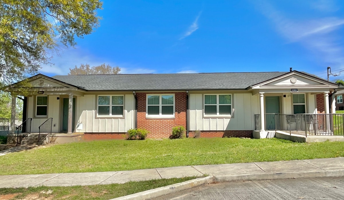 A one-story duplex with a brick and white facade, two front doors, and windows. It is surrounded by a well-kept lawn and framed by trees under a clear blue sky.