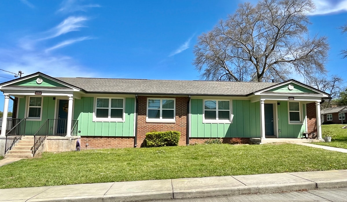 A single-story building with a green and brick facade stands on a sunny day. It features three doors, each with a small set of steps leading up. A neatly maintained lawn and a large tree without leaves are in the background under a clear blue sky.