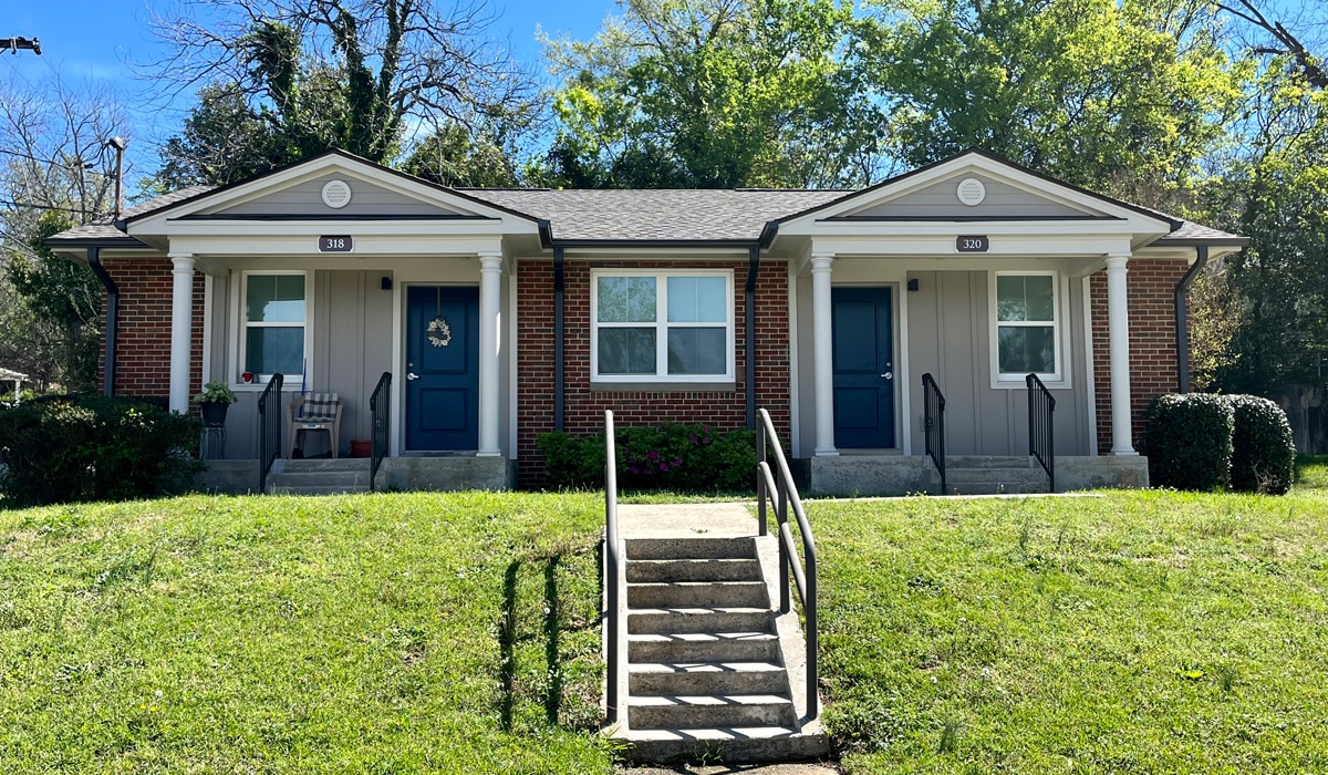 A duplex with brick facade and two blue doors, each with a small front porch. Two sets of stairs and railings lead to the entrances. The building is surrounded by greenery and trees under a clear blue sky.