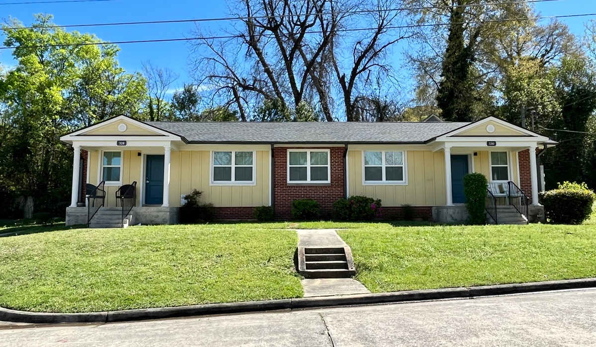 A small brick and beige duplex with two front doors and steps leading to each entrance. The building is surrounded by green grass and trees, with a clear blue sky in the background.