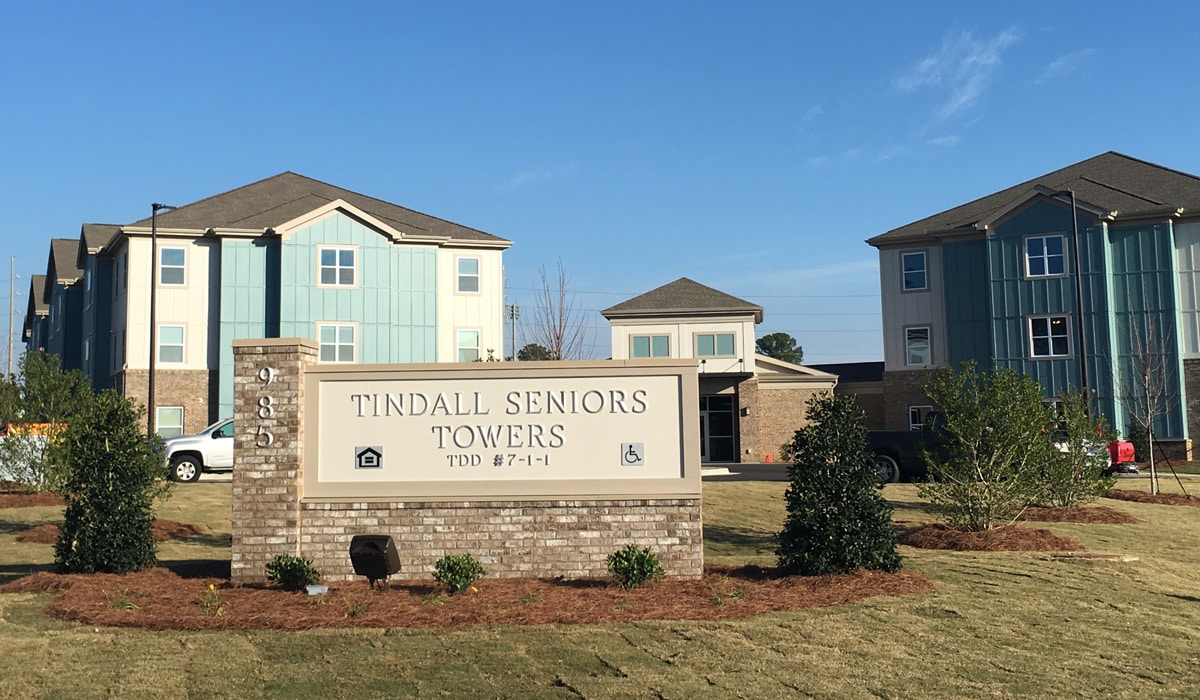 Two modern residential buildings with light blue and brick exteriors stand under a clear blue sky. A sign in the foreground reads 