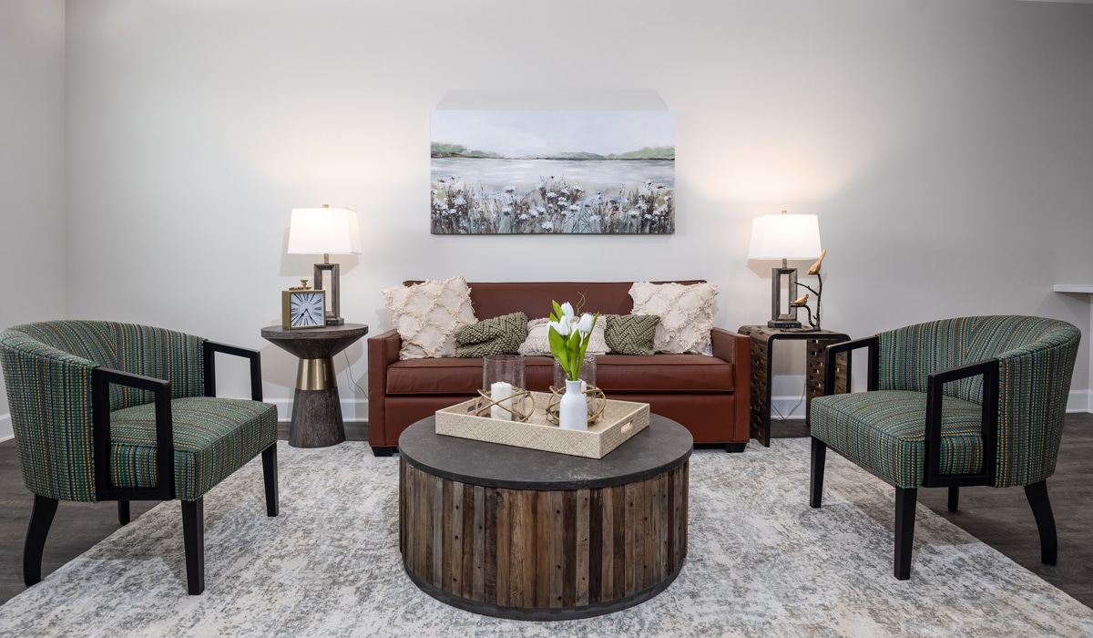 A cozy living room with a brown leather sofa, two green striped armchairs, and a round wooden coffee table. The table holds a decorative tray with flowers. Two lamps flank the sofa, and a landscape painting hangs on the wall.