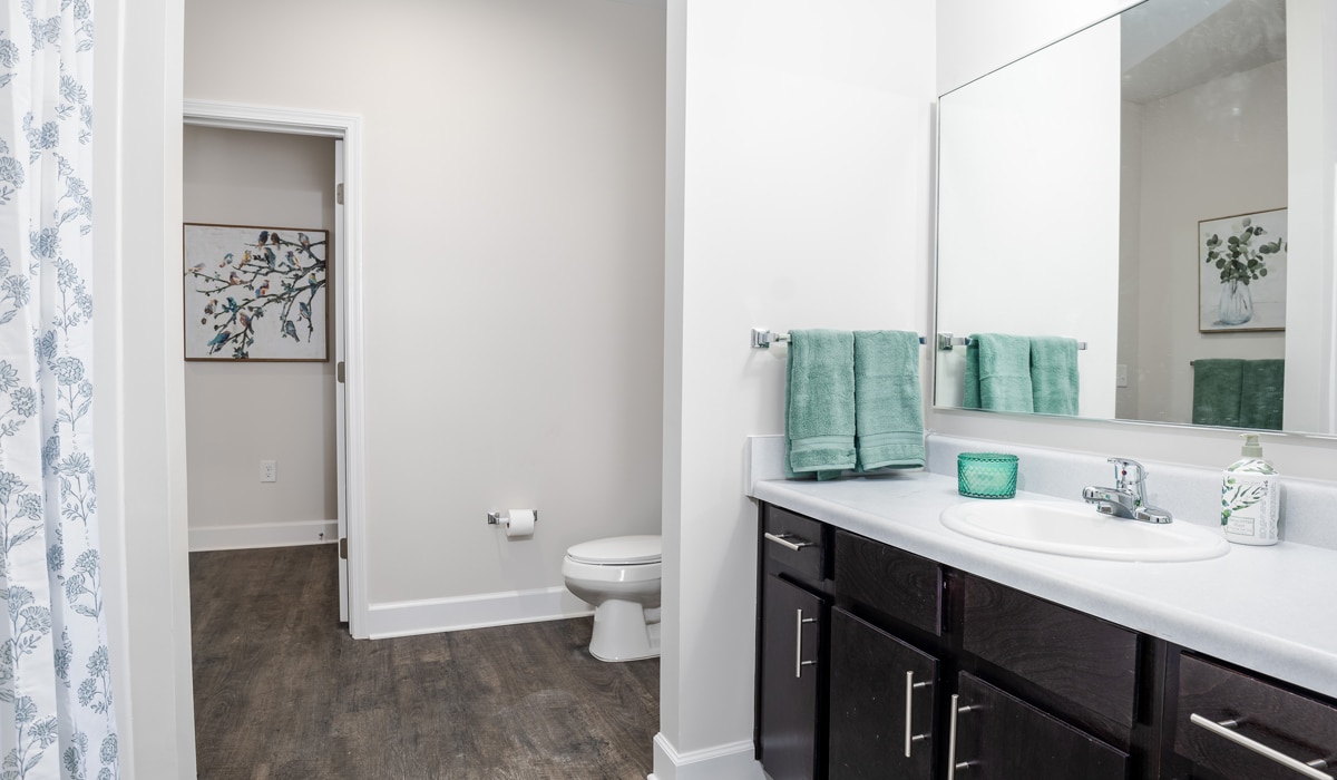 A modern bathroom with dark wood cabinets and a white countertop. There are green towels and a candle near the sink. The toilet is in a separate alcove. A decorative painting hangs on the wall, and the floor is wood-toned.