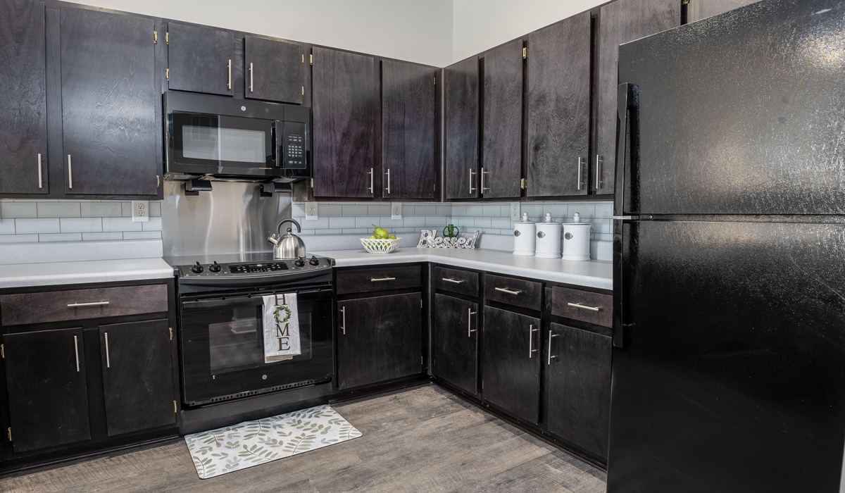 A modern kitchen with dark wooden cabinets, white countertops, a black refrigerator, stove, and microwave. A 