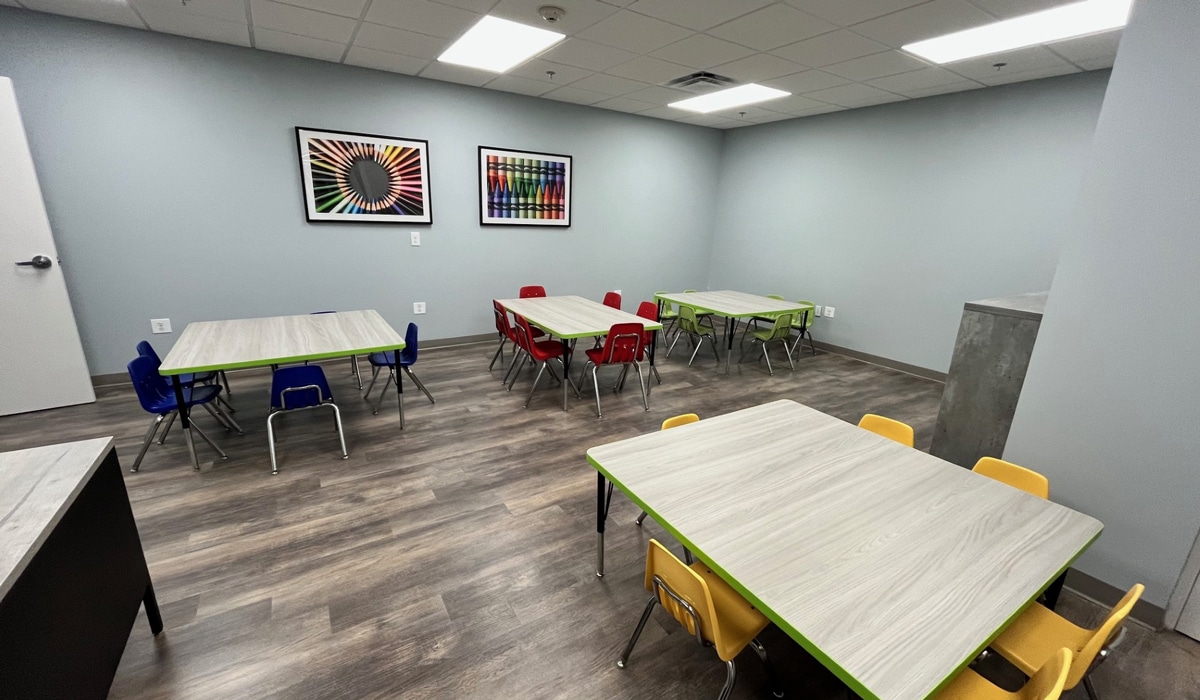 A classroom with three small tables, each surrounded by colorful chairs. Two vibrant, geometric artworks hang on the gray walls. The floor is wooden, and the room is well-lit with overhead lighting.