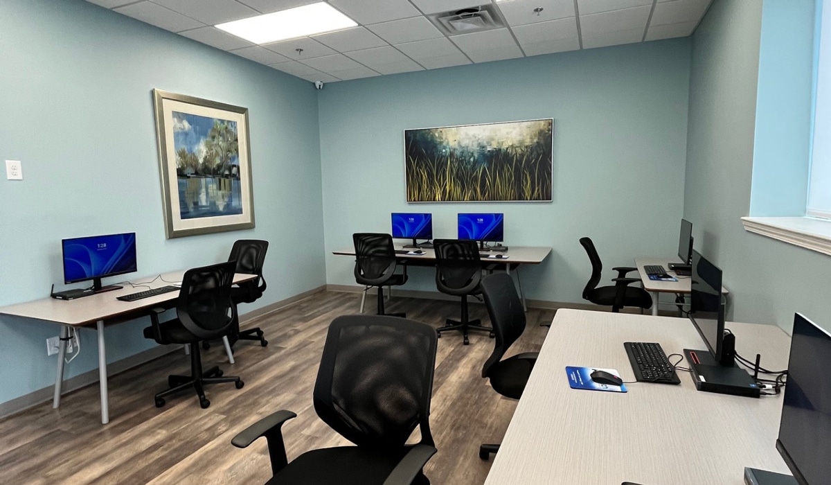 A modern office space with several desks, each equipped with computers and ergonomic chairs. The room has light blue walls, two pieces of framed art, and a large window on the right. The floor is a wood-like material.