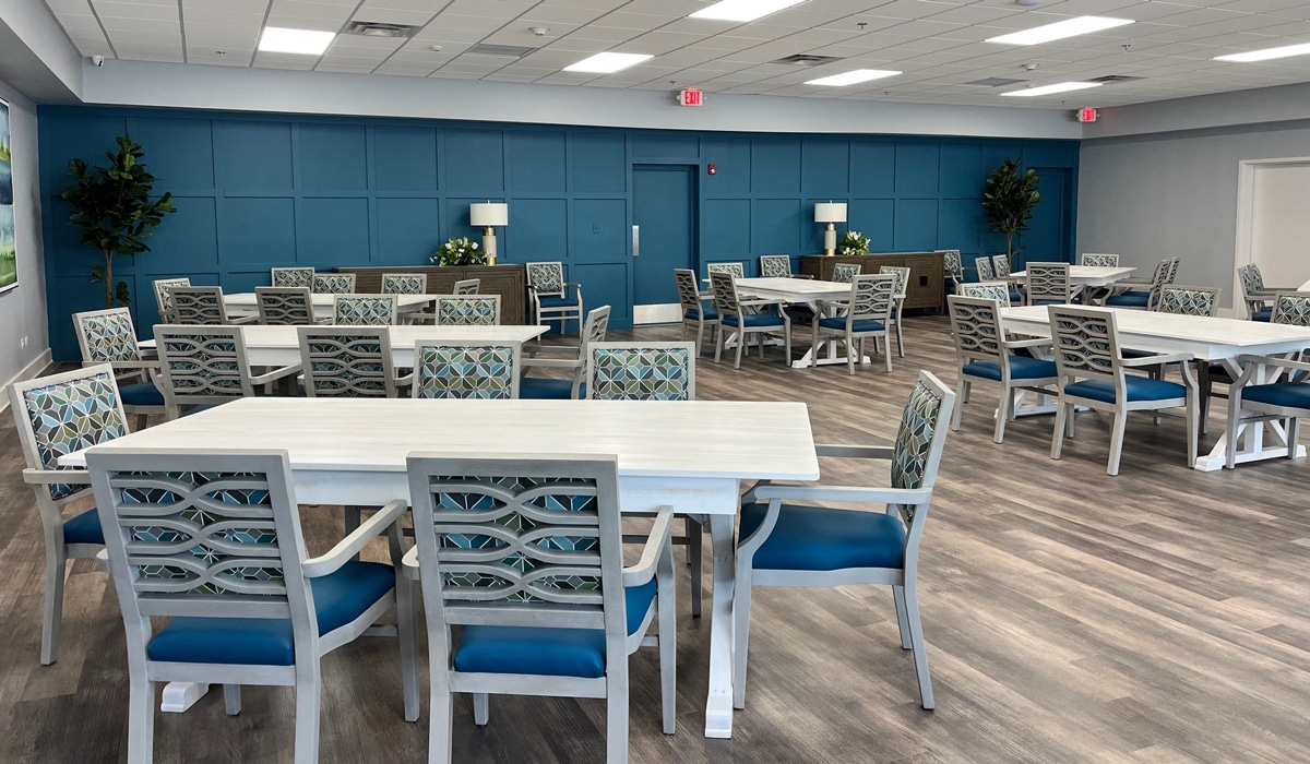 A modern, spacious dining area with several white tables and blue-cushioned chairs. The room features a blue accent wall, wood flooring, and is decorated with potted plants. The space is well-lit with ceiling lights.