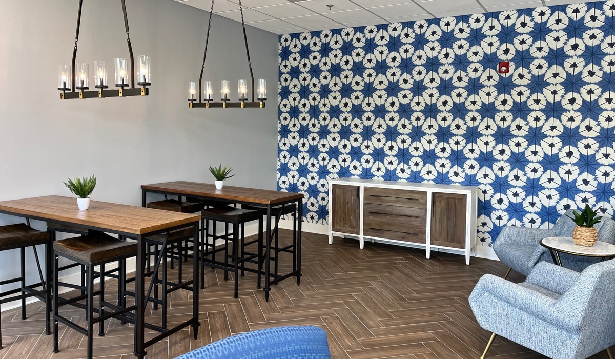 A modern room featuring two wooden tables with black metal stools, a blue and white patterned accent wall, a wooden sideboard, a chandelier with candle-style lights, and two blue upholstered armchairs on a herringbone-patterned floor.