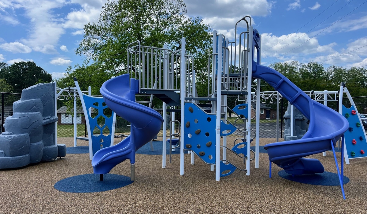 A playground featuring blue slides, climbing structures, and rock walls. The sky is partly cloudy, and trees surround the area. The playground is set on a soft, rubberized surface, providing a safe play environment.