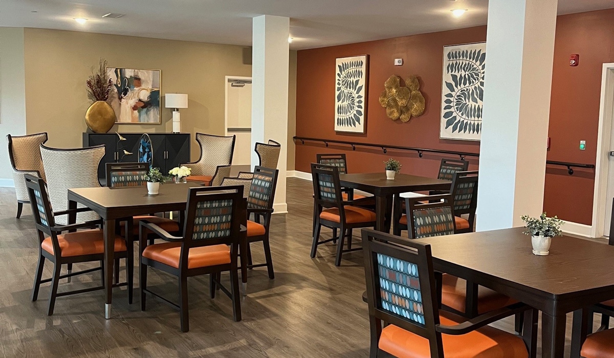 A modern dining area with several wooden tables and chairs featuring orange cushions. The walls are decorated with abstract art and a sculpture. Two tall chairs and a lamp are visible in the corner of the room.